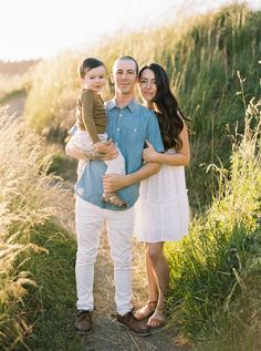 a man and woman holding a baby in their arms while standing on a path surrounded by tall grass