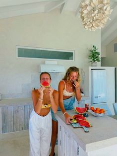 two women standing at a kitchen counter eating watermelon and holding up slices of fruit