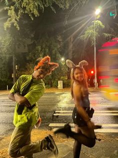 two people in costumes are crossing the street at night, one is dancing and the other is running