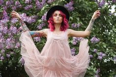 a woman with pink hair wearing a dress and hat standing in front of purple flowers