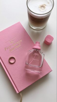 a bottle of perfume sitting on top of a pink book next to a glass filled with liquid