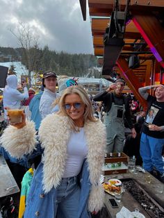 a woman in white shirt and jean pants holding up two glasses with people behind her