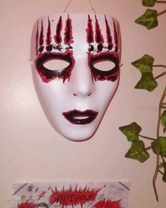 a white mask with red paint on it hanging from a wall next to a plant