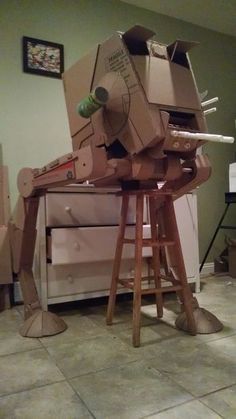 a robot made out of cardboard sitting on top of a wooden stool in a room