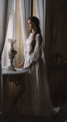 a woman standing in front of a window next to a table with a vase on it