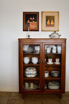 a wooden cabinet filled with dishes and plates on top of a tiled floor next to two framed pictures