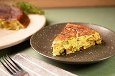 a slice of quiche sits on a plate next to a fork and knife, along with another dish