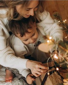 a woman and her son are looking at a present under the christmas tree with lights