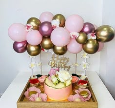 a birthday cake with pink, gold and white balloons above it on a wooden box