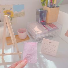 a hand holding a piece of pink tissue next to a desk with art supplies on it