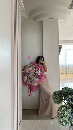 a woman in a long dress holding a bouquet of flowers while standing next to a wall