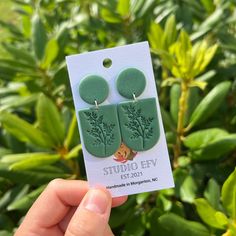 a person holding up a pair of green earrings in front of some bushes and trees