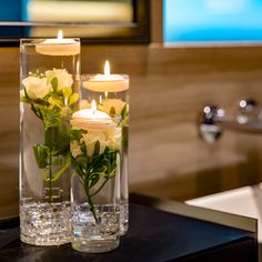 two clear vases filled with white roses and lit candles on a black table in a bathroom