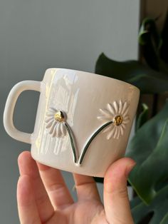 a hand holding a white coffee mug with two flowers on the outside and gold centers