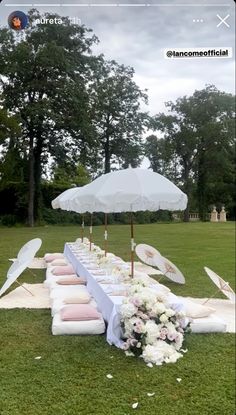 an outdoor table set up with umbrellas and flowers on the grass in front of it