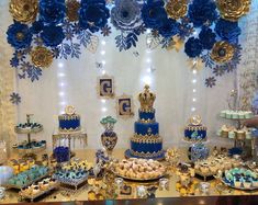 a table topped with lots of blue and gold desserts next to a wall covered in paper flowers