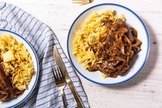 two plates with pasta and meat on them next to a fork, knife and napkin