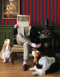 a man sitting in a chair reading a newspaper with two dogs watching him and another dog laying on the floor