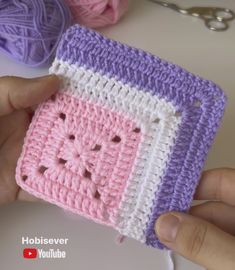 a hand holding a crocheted square with holes in the middle and two balls of yarn next to it