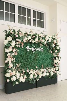 a green wall with white flowers and greenery on the top is surrounded by black planters