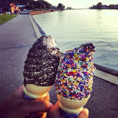 someone holding two ice cream cones with sprinkles on them near the water