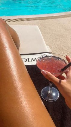 a woman holding a drink in her hand next to a swimming pool with the sun shining on her legs