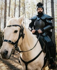 a man in armor riding on the back of a white horse next to a forest