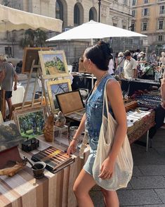 a woman standing next to a table with paintings on it and an umbrella in the background