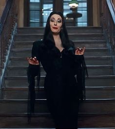 a woman in black dress standing on stairs with her hands out and looking at the camera