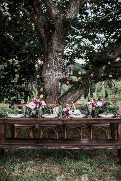 a wooden table with plates and flowers on it in front of a large oak tree