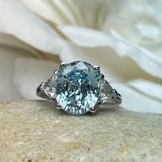 "The ring pictured is simulated diamond aquamarine #7126 -Approximate total carat weight: 5.00ctw diamond equivalent -Center Stone Size: 11x9mm - approx. 4.20ct diamond equivalent -Center Stone Shape: oval cut -Side Stones Size: 2 Trillions 5x5mm - approximately 0.80ctw diamond equivalent -Gem Type: simulated diamond -Stone Clarity: VVS1 -Stone Color: Blue Aquamarine -Moh's Scale: 8.5 hardness -Metal Type and Purity: 14k white gold -Setting: 4 prong tulip head -Stock Ring Size: 6 -Country of Man Trillion Engagement Ring, Engagement Ring Three Stone, Ring Three Stone, Blue Sapphire Necklace, Aquamarine Engagement Ring, White Gold Set, Oval Ring, Ring Pictures, Diamond Wedding Ring