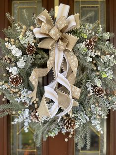 a christmas wreath with pine cones and white ribbon hanging on the front door to welcome guests