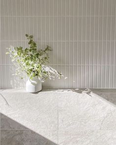 a white vase filled with green plants sitting on top of a tiled floor next to a wall