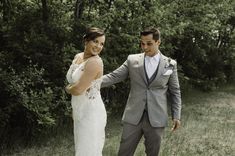 a bride and groom standing in the grass near some trees, smiling at each other