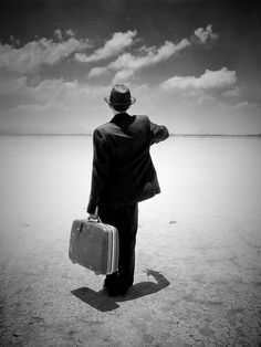 a man in a suit and hat carrying a suitcase on the desert plain under a cloudy sky