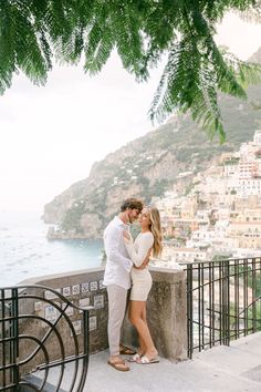 a man and woman standing next to each other on a balcony near the ocean with buildings in the background