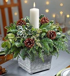 a white candle is sitting on top of a silver container filled with greenery and pine cones