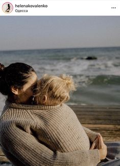 a woman sitting on top of a wooden bench next to the ocean with her hair in a bun