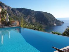 an empty swimming pool overlooking the ocean and mountains with chairs around it, on a sunny day