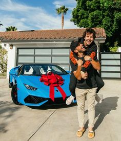 a man carrying a woman in front of a blue sports car with a red bow on it