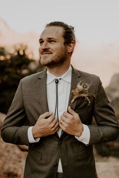 a man wearing a suit and tie standing in front of some mountains with his hands on his chest