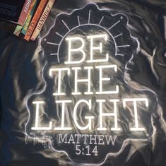 a light up sign that says be the light with books in the back ground behind it