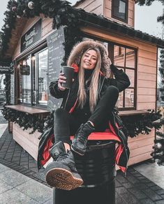 a woman sitting on top of a trash can next to a small building with christmas decorations