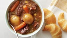 a bowl of beef stew and croissants on a white cloth with a spoon