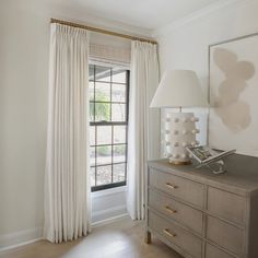 a bedroom with a dresser, lamp and large window in the corner that has white drapes on it