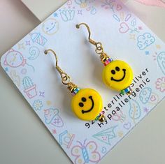 two yellow smiley face earrings are on a card with a pink and blue ribbon around it