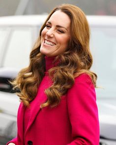 a woman in a pink coat smiles as she stands next to a white car and looks off into the distance