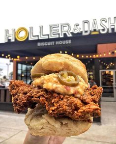 a person holding up a large sandwich in front of a building that says holler & dash biscuit house