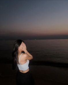 a woman standing on top of a beach next to the ocean at night with her hands covering her face