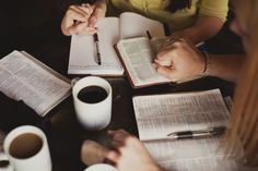 three people are sitting at a table with books and cups of coffee, one is writing